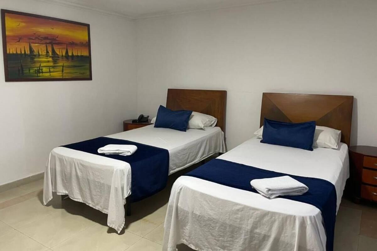 Quarto de hotel com duas camas de solteiro, cada uma com cabeceira de madeira, roupa de cama branca e detalhes azuis. Um quadro de um pôr do sol está na parede.