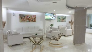 A modern, well-lit lobby with white sofas, a coffee table, and two rocking chairs. There are two large paintings on the walls and a flat-screen TV mounted above a seating area. - Hotel Charthon (Barranquilla)