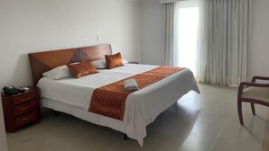 A hotel room with a large bed, a wooden headboard, a bedside table with a phone, and a chair. - Hotel Charthon (Barranquilla)