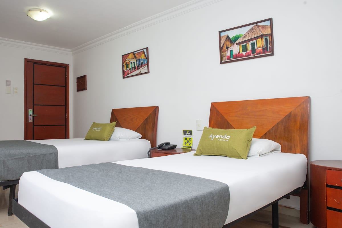 Quarto de hotel com duas camas, cada uma com cabeceira de madeira, roupa de cama branca e almofada verde. Apresenta uma mesa de cabeceira de madeira com telefone e uma pequena mesa com armário vermelho.