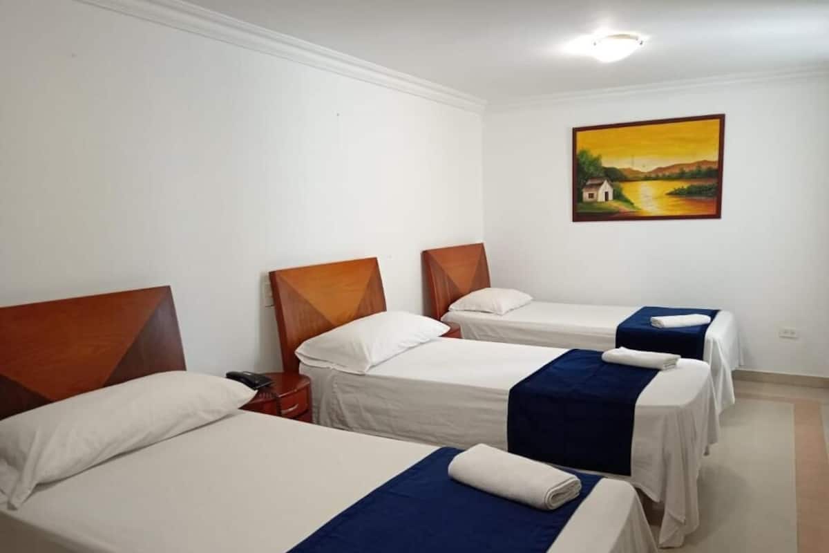 Quarto de hotel com três camas, cada uma com cabeceira de madeira e roupa de cama branca. Há uma pintura na parede retratando uma paisagem.