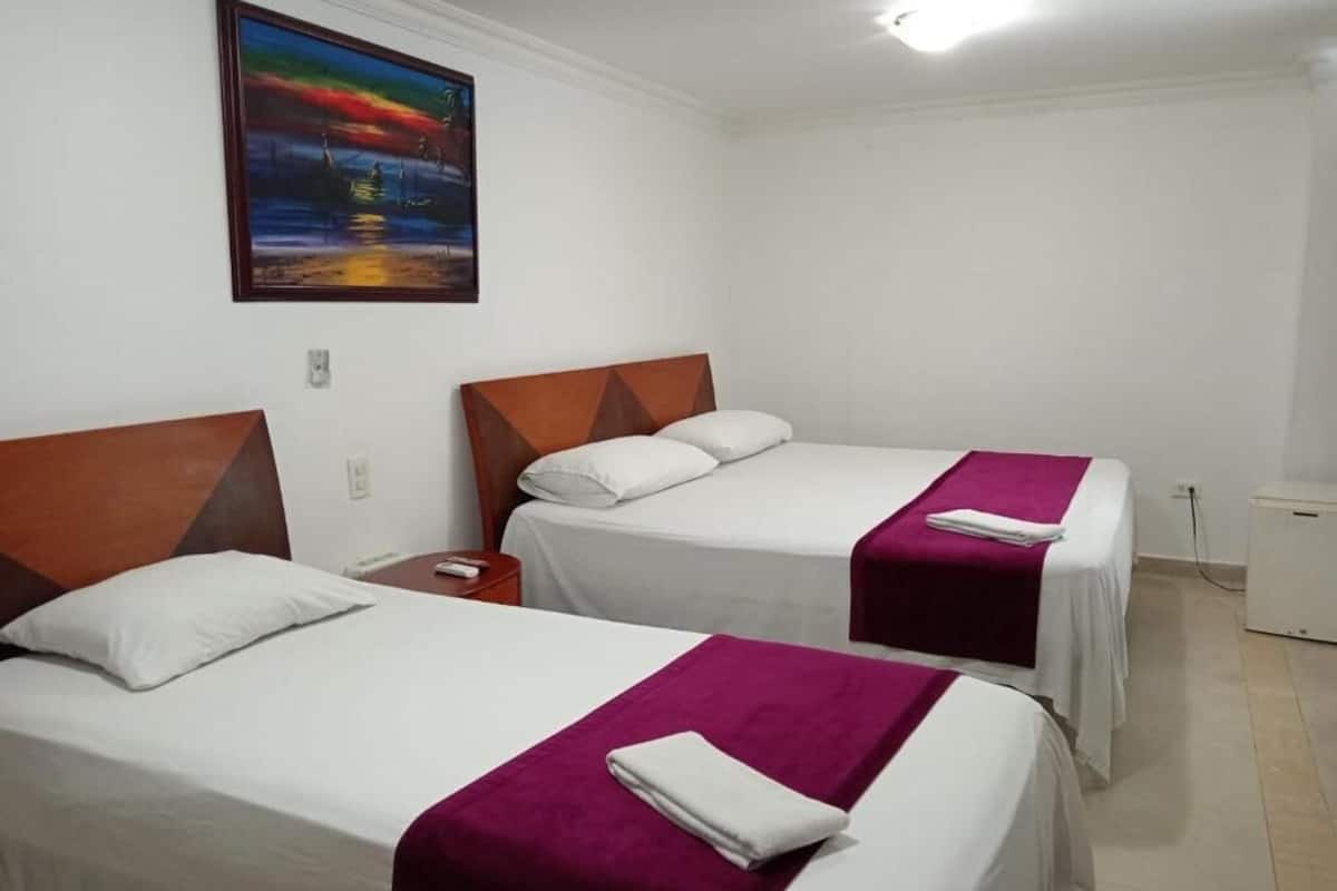 Quarto de hotel com duas camas, cada uma com cabeceira de madeira, roupa de cama branca e manta roxa. Há um quadro na parede acima das camas.