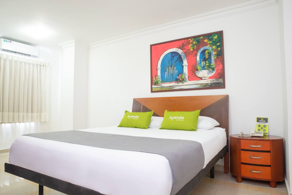 Quarto de hotel com cama, mesa de cabeceira, quadro emoldurado e janela com cortinas.