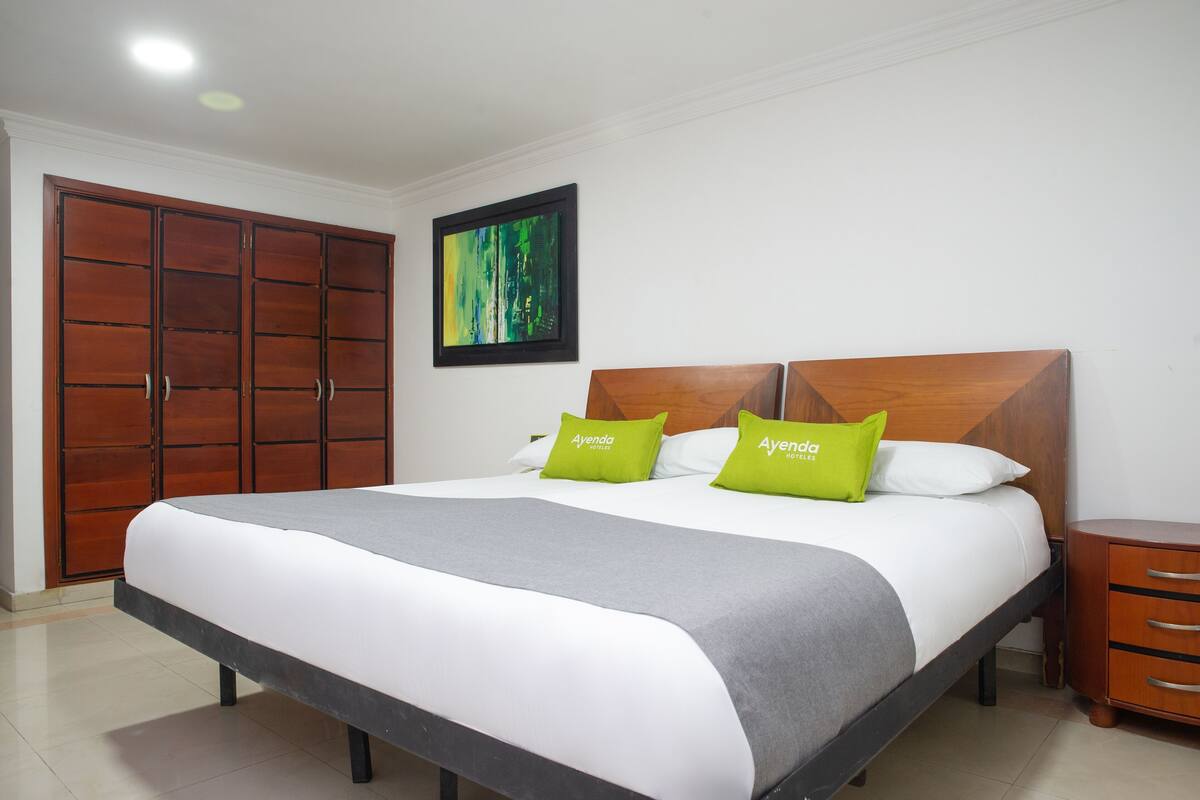 Quarto com cama, duas almofadas, cabeceira de madeira, mesa de cabeceira, guarda-roupa de madeira e um quadro na parede.