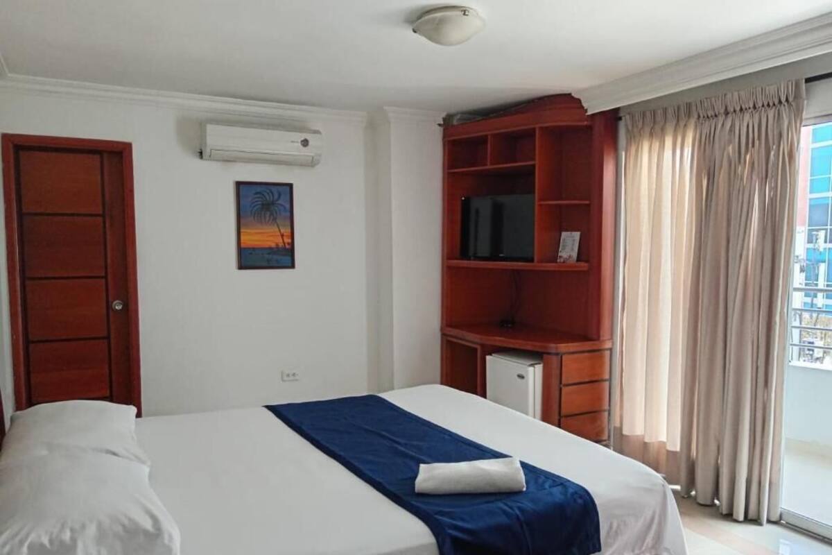 Quarto de hotel com cama, televisão sobre um móvel de madeira e janela com cortinas.