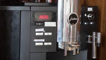 Una cafetera con una taza de café sobre un platillo que dice "HORNIG KAFFEE" y un recipiente con granos de café.