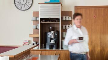 Una zona de cocina con cafetera, estantes con platos y tazas, y un reloj en la pared.