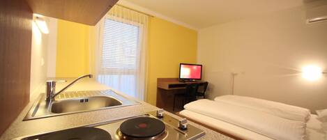 Habitación de hotel con kitchenette, cama, escritorio con computadora y ventana con persianas.
