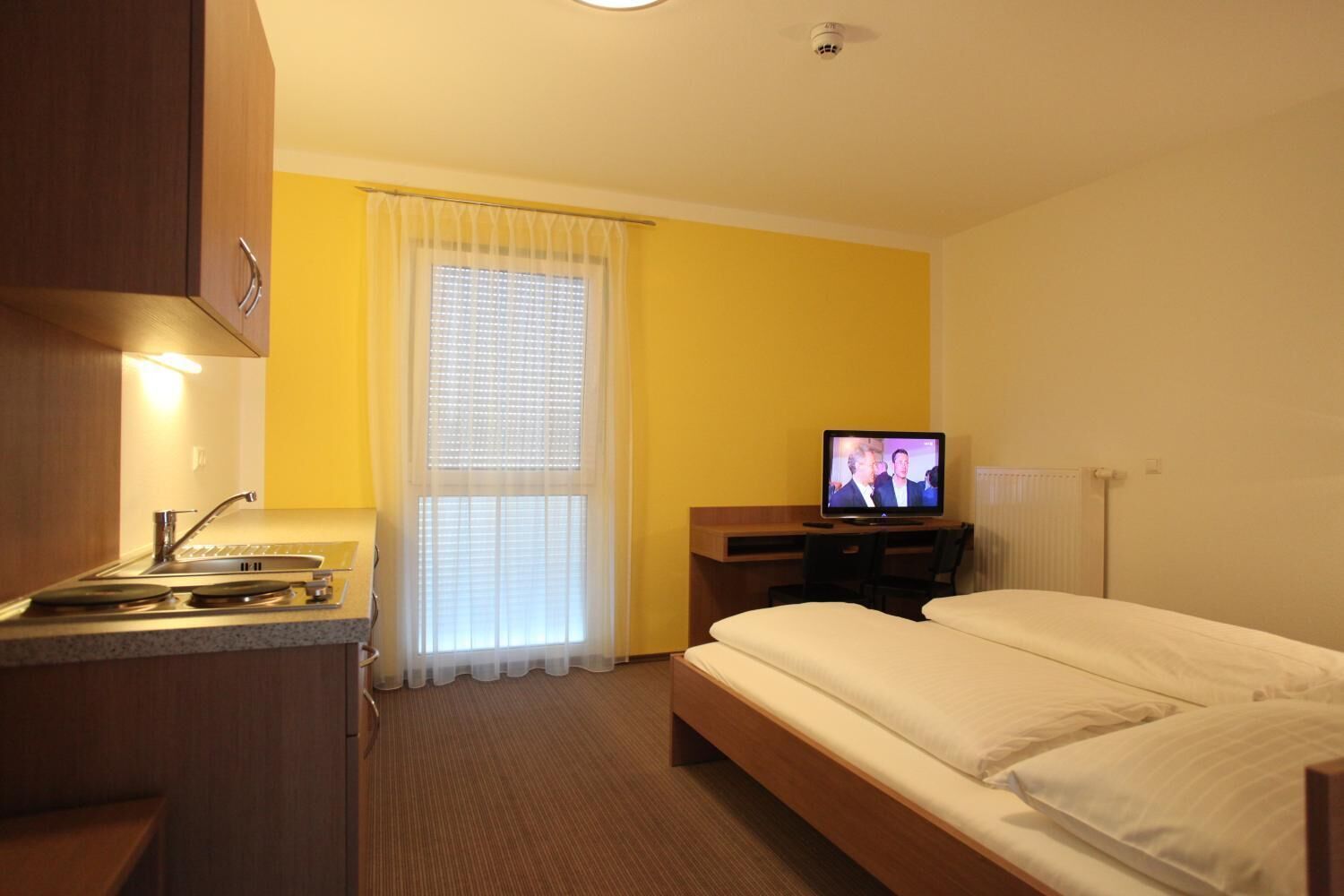 Habitación de hotel con cama, escritorio con televisión, ventana con persianas y kitchenette.