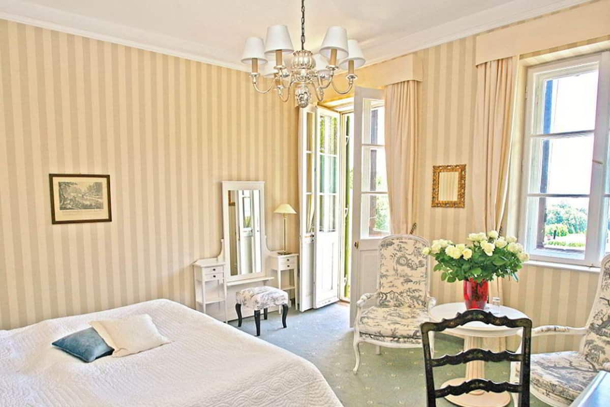 A hotel room with a large bed, a sitting area with a chair and a table, a mirror, and a window with curtains.