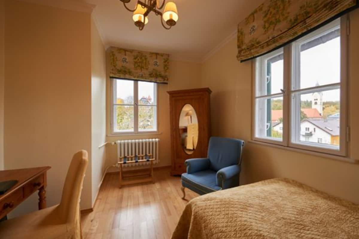 A room with a wooden floor, a blue armchair, a wooden cabinet, a radiator, and a window with a view of houses.