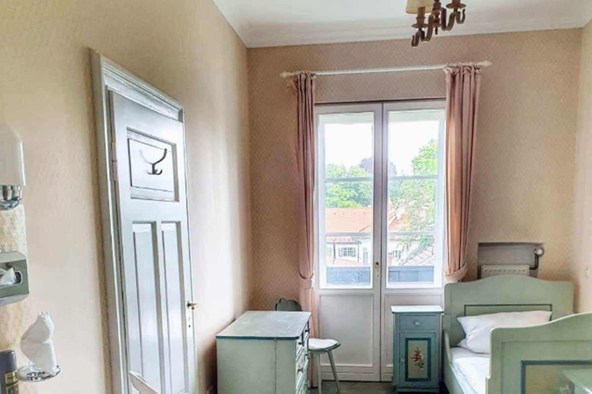 A small, compact room with a bed, a desk, a chair, and a window with curtains.