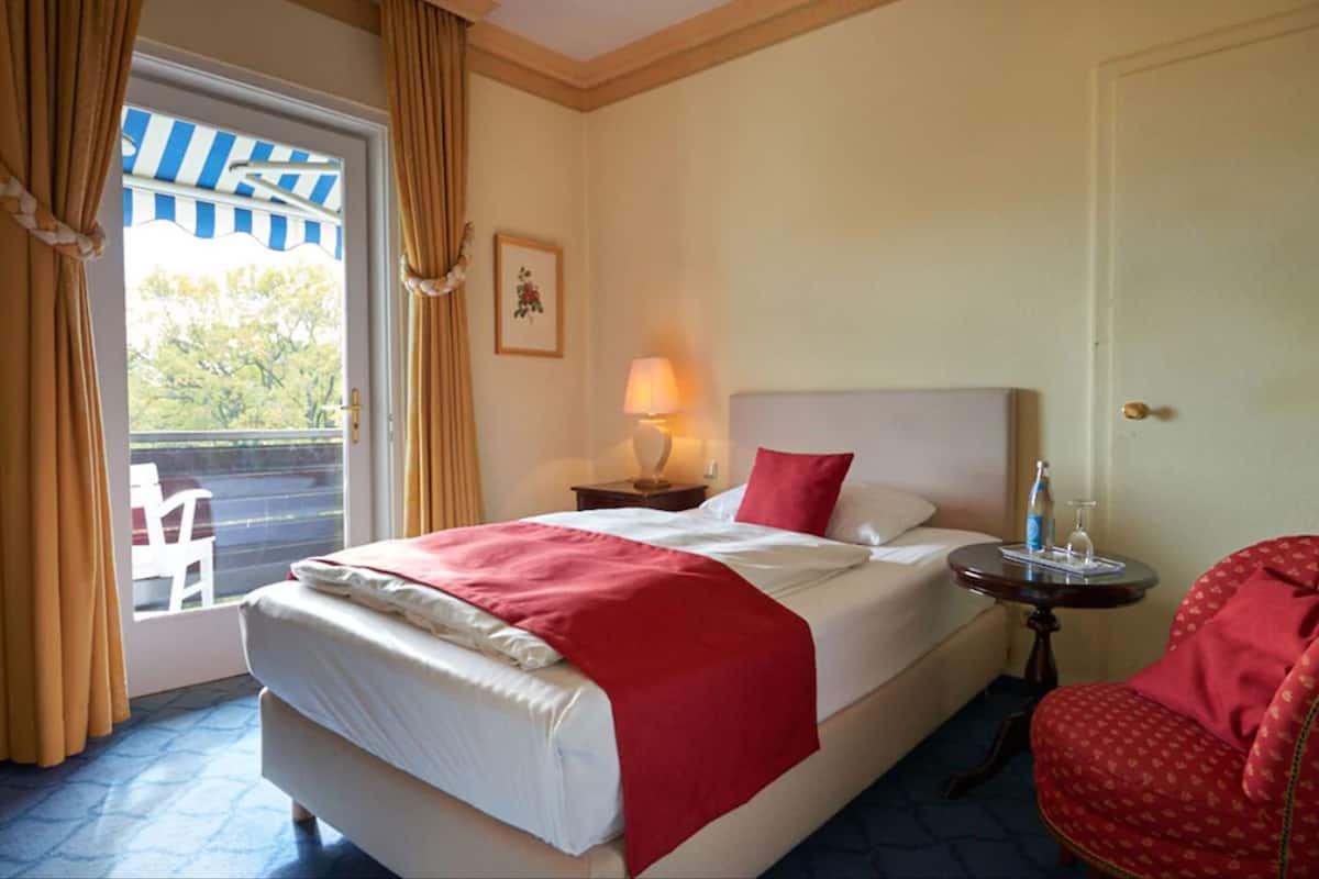 A hotel room with a bed, a chair, a small table with a lamp, and a window with curtains.
