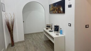 Un espace d’accueil d’hôtel avec un comptoir de réception blanc, un tableau encadré représentant un bâtiment, un panneau indicateur et une plante décorative.