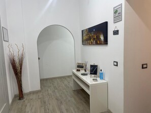 A hotel reception area with a white reception desk, a framed picture of a building, a signboard, and a decorative plant.