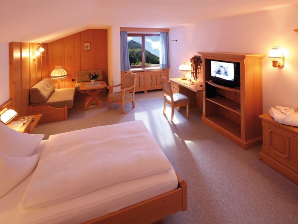 A hotel room with a bed, a desk with a TV, a chair, a small table, and a window with a view of the mountains.