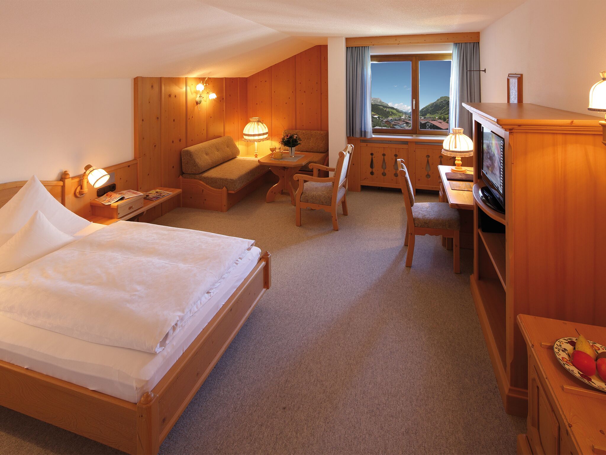 A cozy hotel room with a bed, a sofa, a desk with a television, and a view of the mountains.