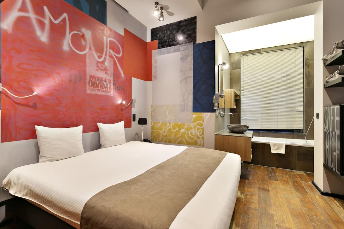 Une chambre d’hôtel moderne avec un grand lit, une salle de bain dotée d’un miroir et d’un lavabo, et une fenêtre avec des stores.