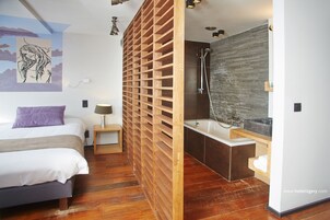 Una habitación de hotel moderna con un separador de madera, una cama grande, una mesita de noche y un baño con ducha y lavabo.