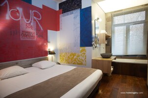 Una habitación de hotel con cama, mesita de noche, lámpara, ventana con persianas y baño con lavabo y espejo.
