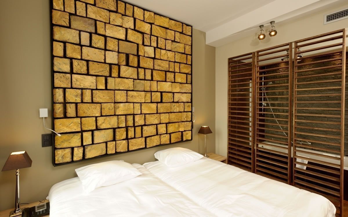 A hotel room with a large brick-patterned wall, a bed with white linens, a nightstand with a lamp, and a wooden wardrobe with horizontal slats.