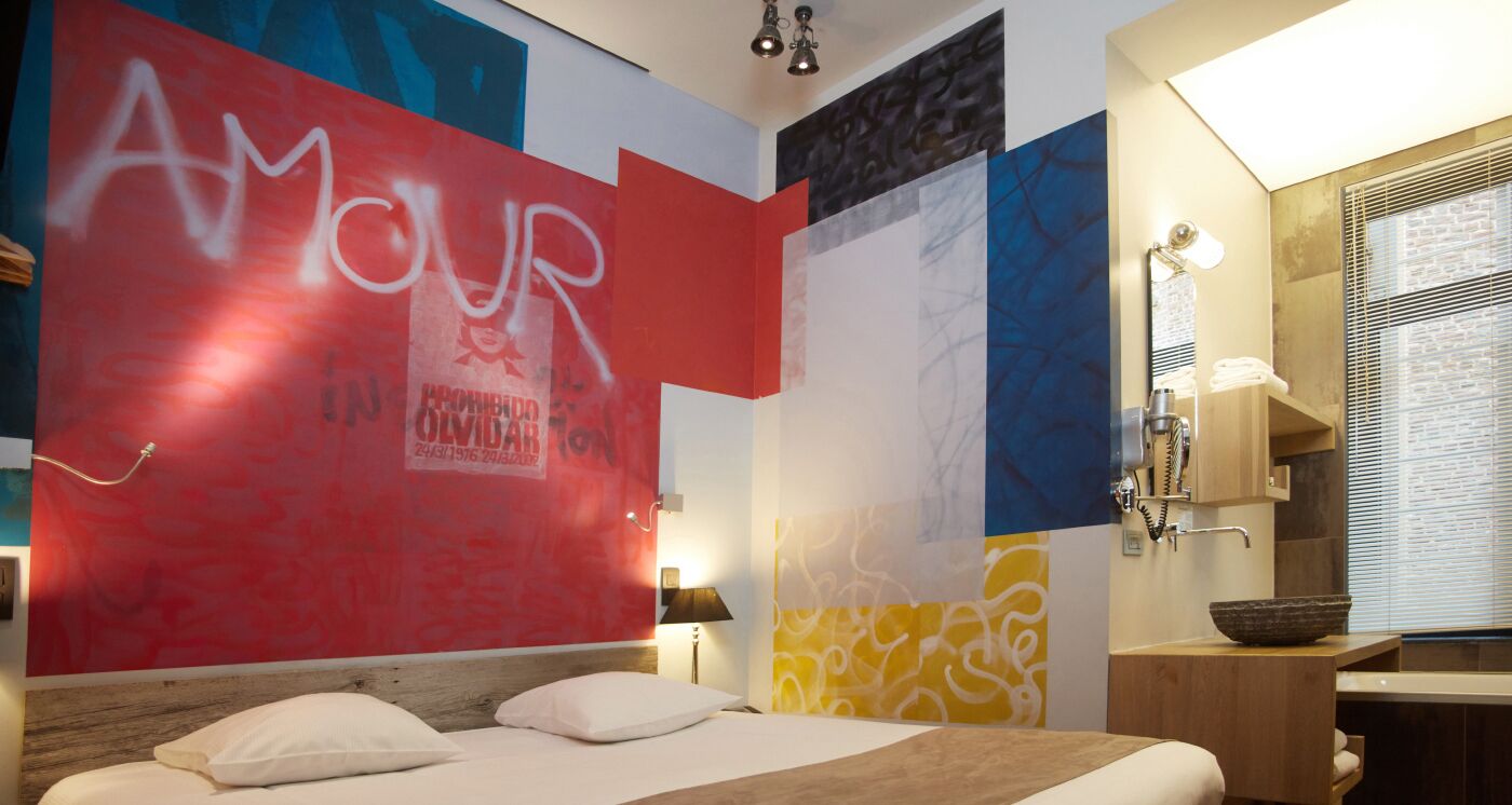 A hotel room with a bed, a bedside table, a mirror, and a bathroom with a sink and a bowl on the counter.