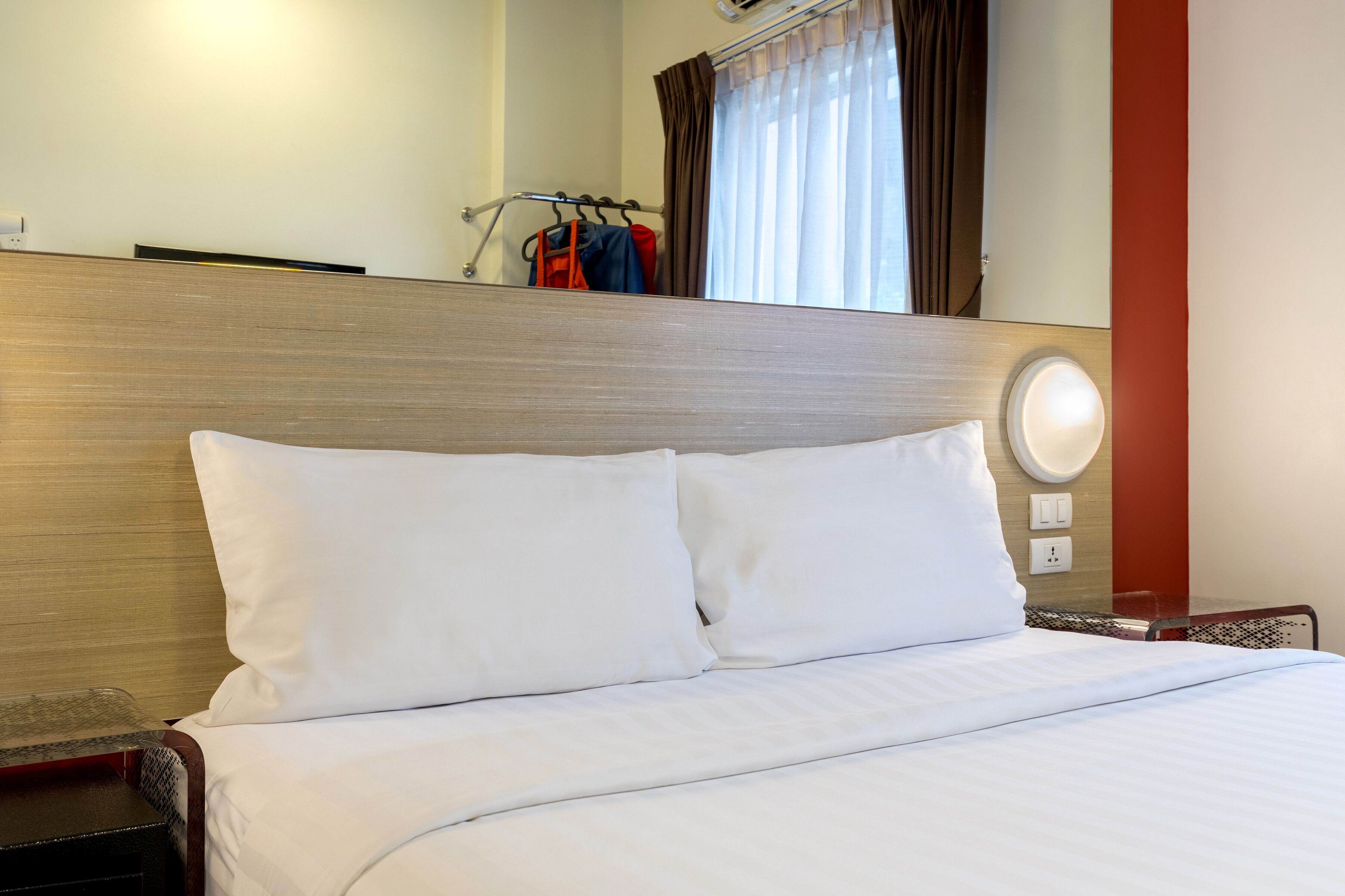 A hotel room with a wooden headboard, white bedding, a bedside table, and a coat rack with clothes.