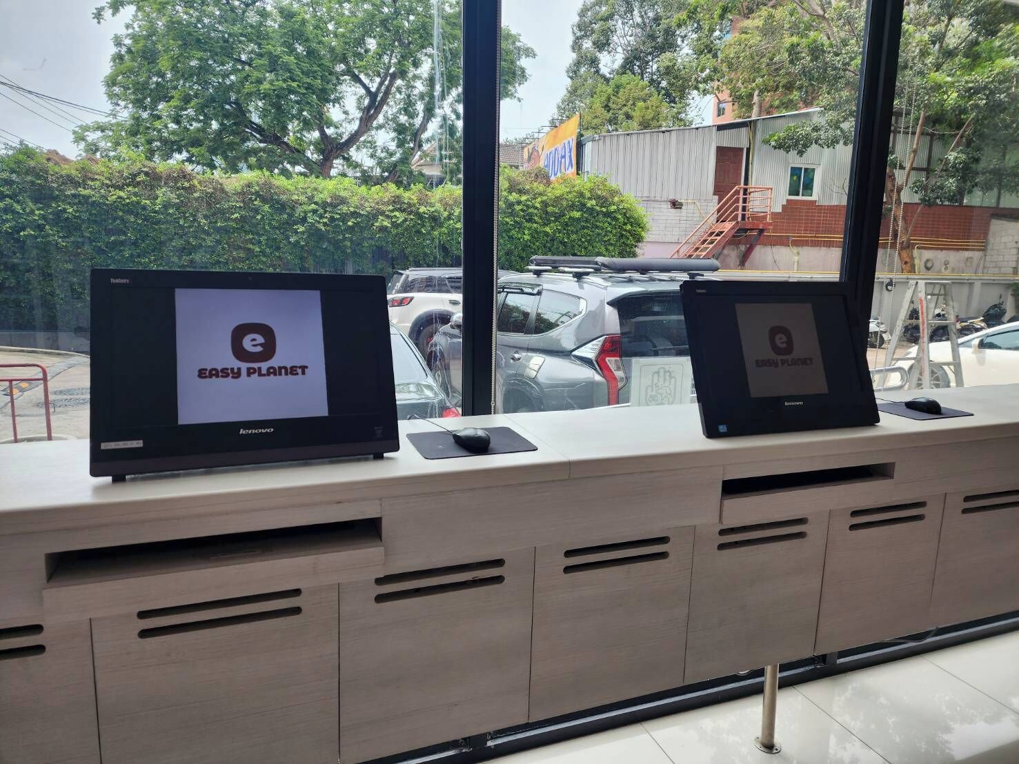 A reception desk with two monitors displaying the 'Easy Planet' logo, a window with a view of parked cars and trees, and a metal railing.