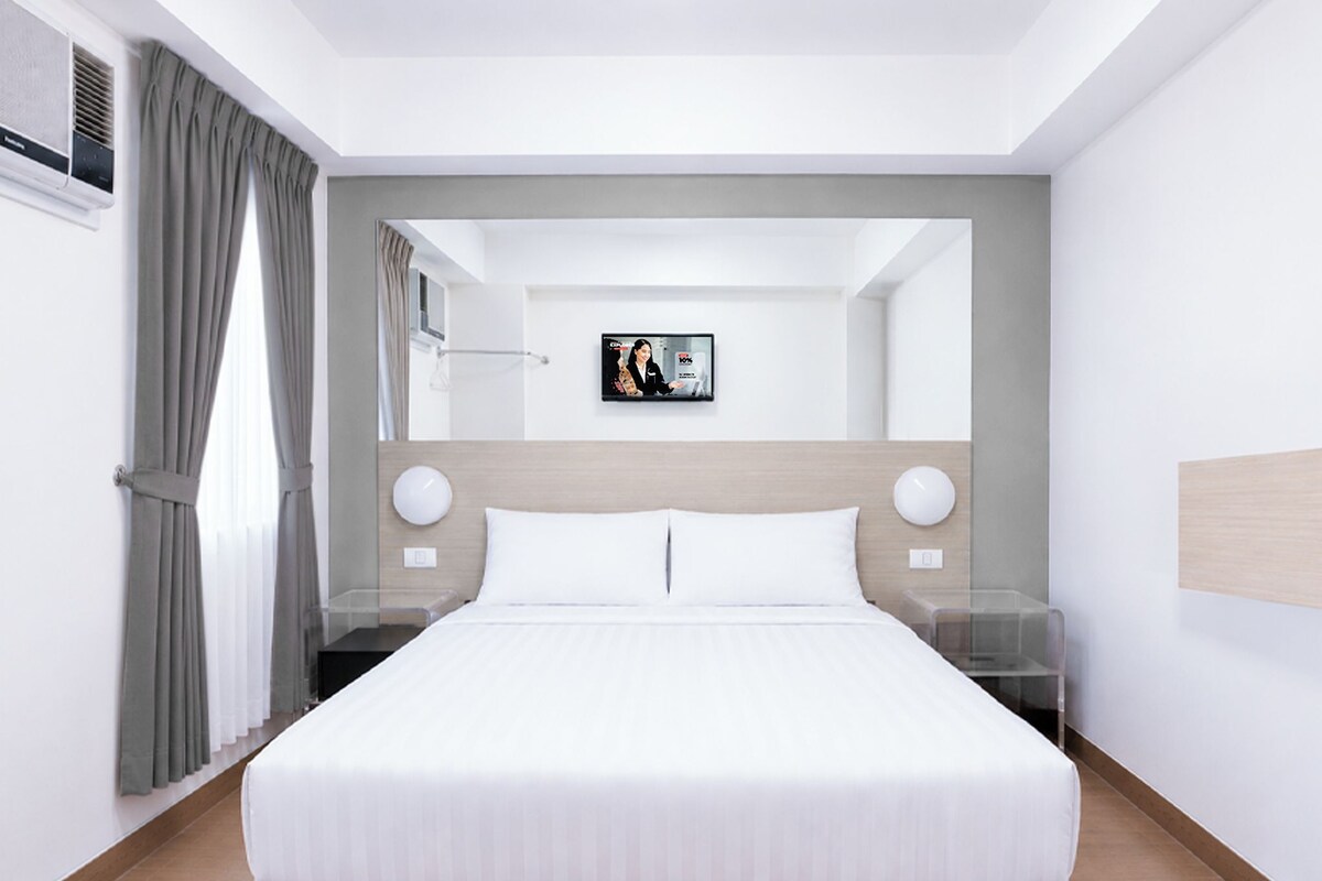 Una habitación de hotel moderna con una cama grande, mesitas de noche, un televisor de pantalla plana y vistas al pasillo.