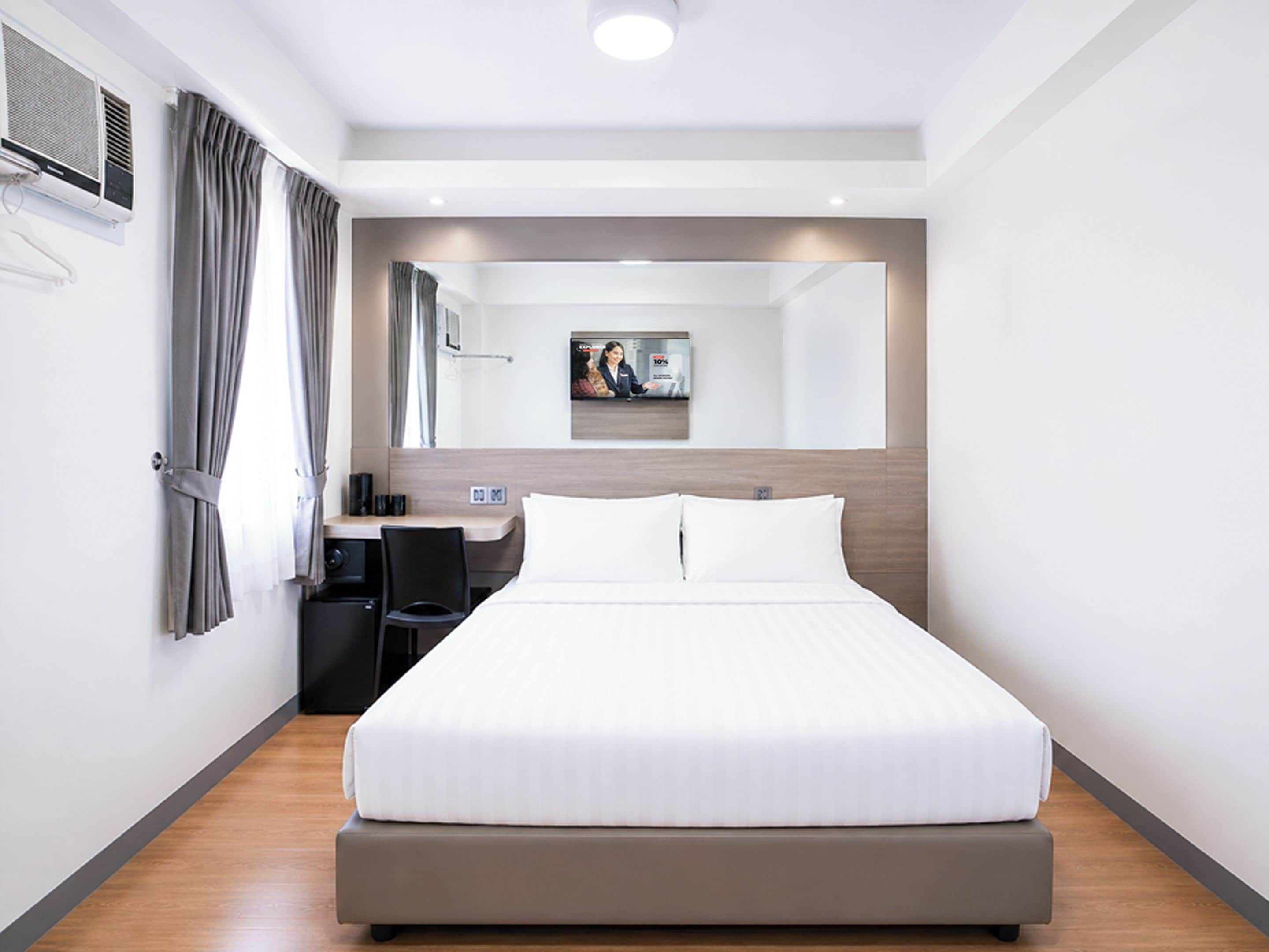 A modern hotel room with a large bed, a desk, a chair, a flat-screen TV, and a mirror.