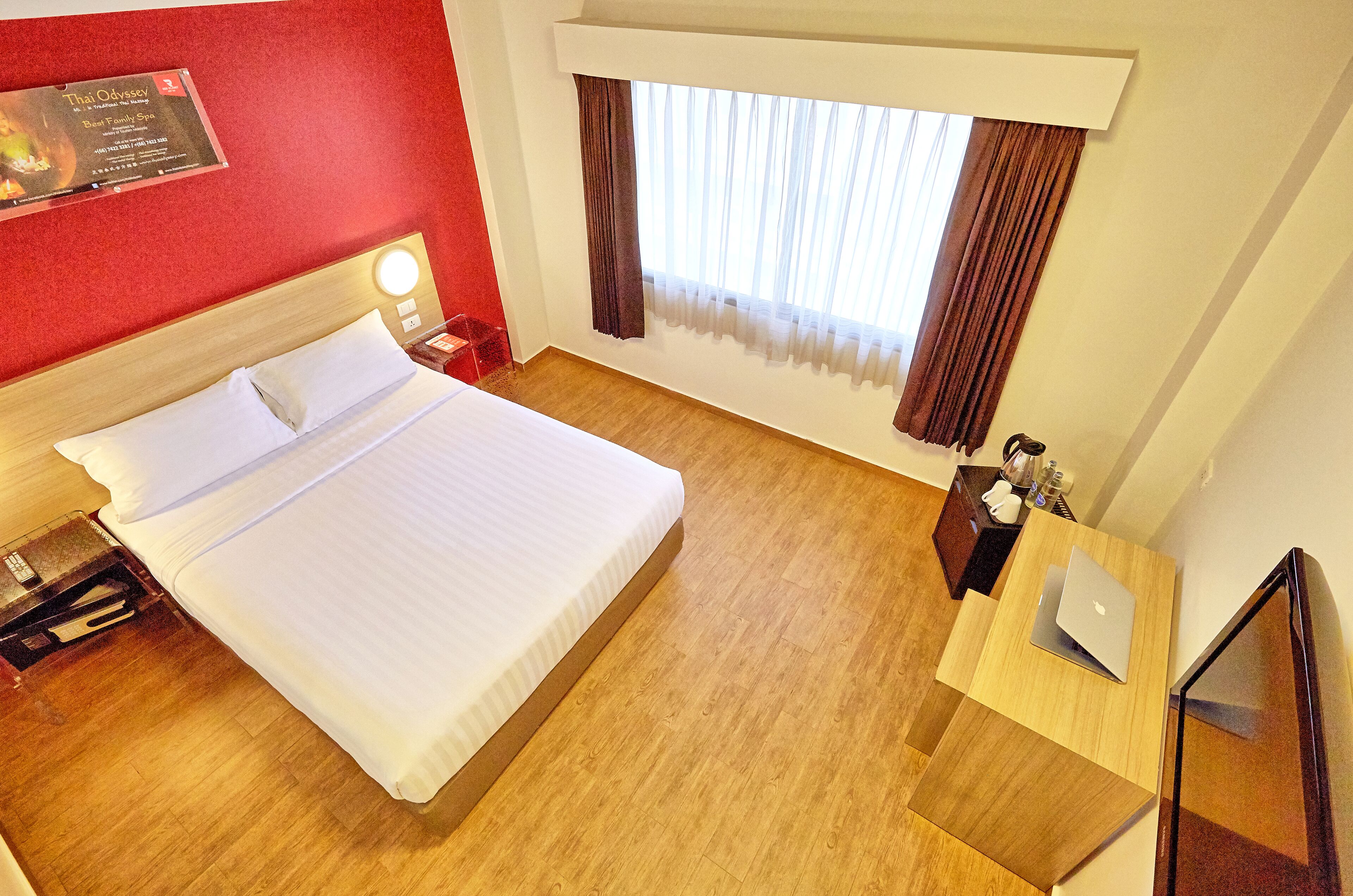 A hotel room with a bed, a desk with a laptop, a window with curtains, and a red wall with a sign.