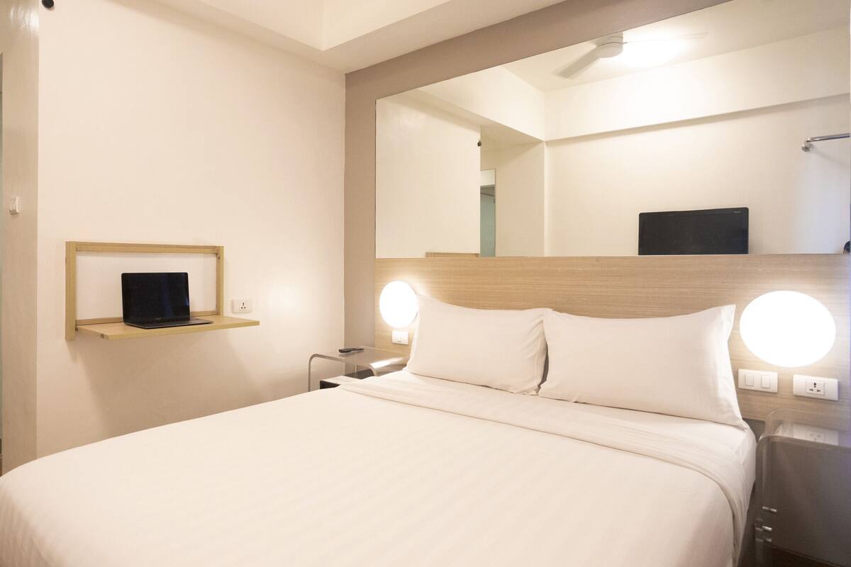 Habitació d'hotel amb un llit, una taula de nit, una làmpada, un mirall i una petita estant amb un ordinador portàtil.