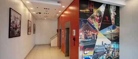 A hotel corridor with a red accent wall, framed pictures on the left, and a large poster with various travel scenes on the right.