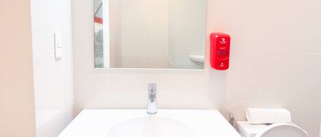 A bathroom with a white sink, a mirror, a red soap dispenser, and a white toilet.