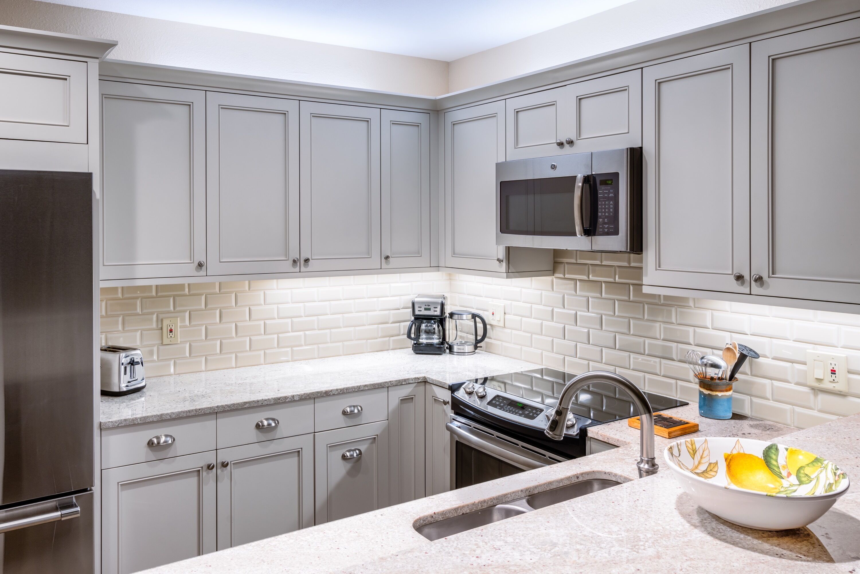 A kitchen with grey cabinets, a stainless steel refrigerator, a microwave, a coffee maker, and a sink with a bowl of lemons.