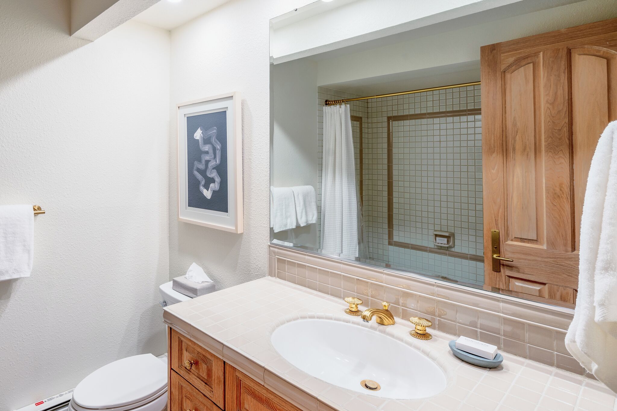 A bathroom with a white sink, wooden vanity, a framed artwork, and a large mirror.