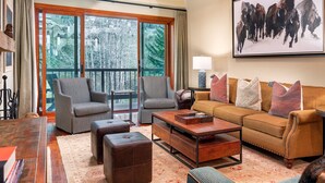 A spacious living room with a large window, a sofa, two armchairs, a coffee table, and a bookshelf.