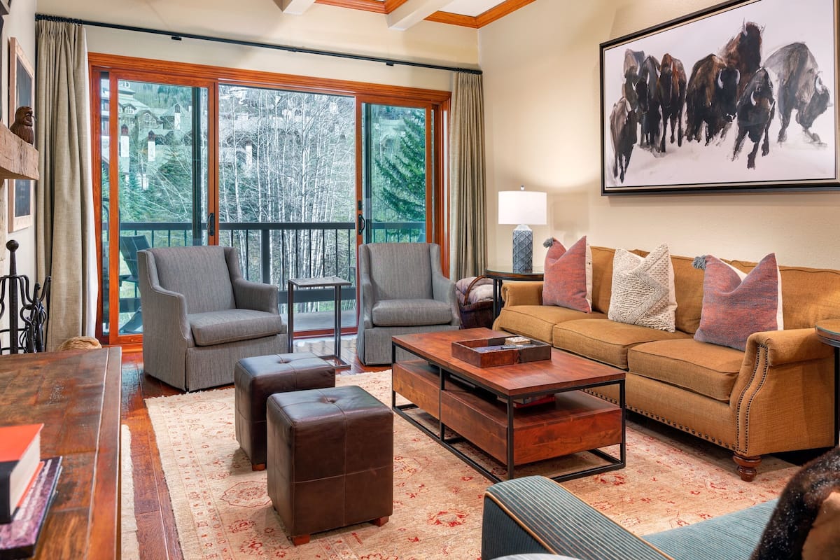 A spacious living room with a large window, a sofa, two armchairs, a coffee table, and a bookshelf.