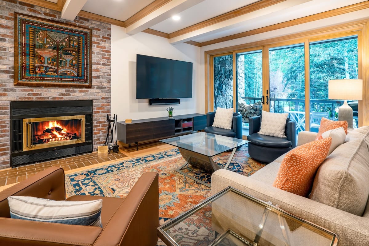 A living room with a fireplace, a flat-screen TV, and a large window with a view of trees.