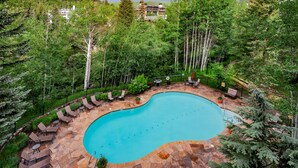 A swimming pool surrounded by lounge chairs and potted plants, with a view of a house and forested area in the background.