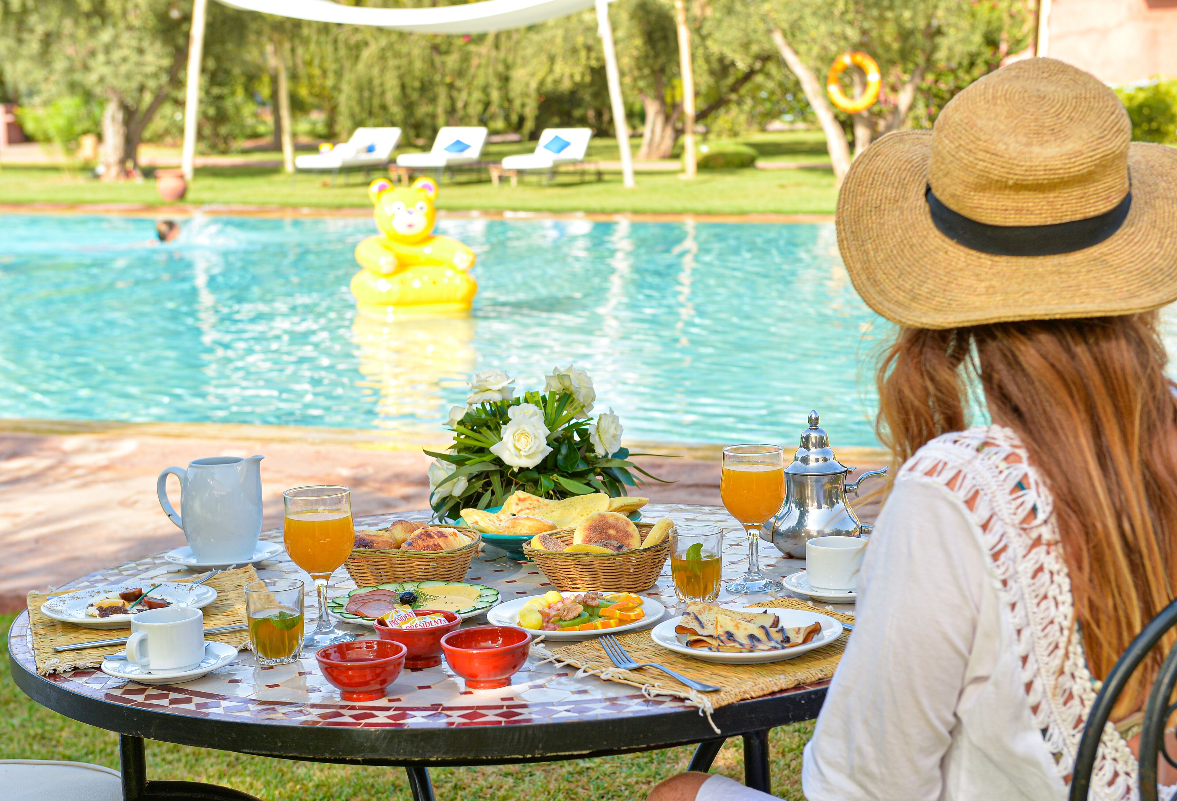 A dining table with various food items and drinks, a pool with a yellow inflatable bear, and a person wearing a straw hat.