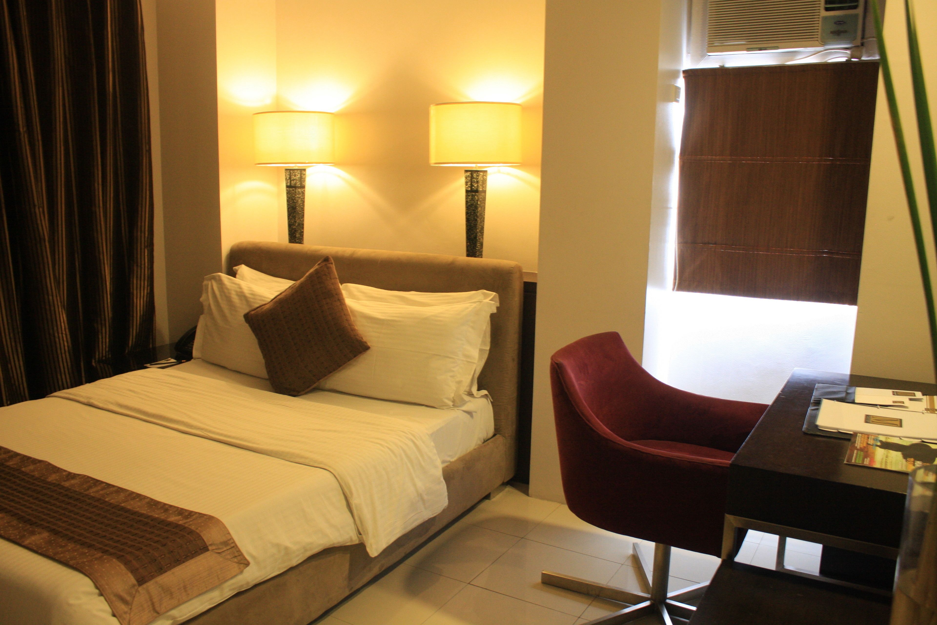 Habitación de hotel con una cama, dos lámparas de noche, una silla roja y un escritorio con lámpara.