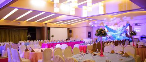 A banquet hall with round tables, white chairs, and a stage with draped fabric and balloons.