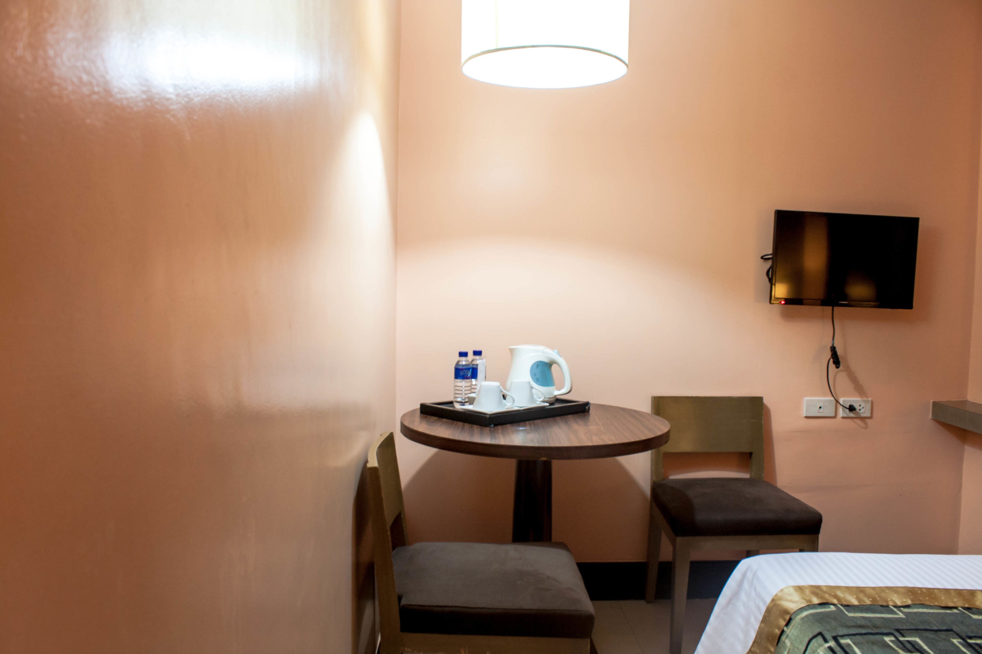 Ein Hotelzimmer mit einem Bett, einem kleinen runden Tisch mit Wasserkocher und Wasserflaschen, einem Stuhl und einem an der Wand befestigten Fernseher.