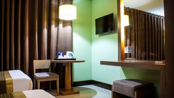A hotel room with a bed, a chair, a small table with a lamp, a TV mounted on the wall, and a window with curtains.