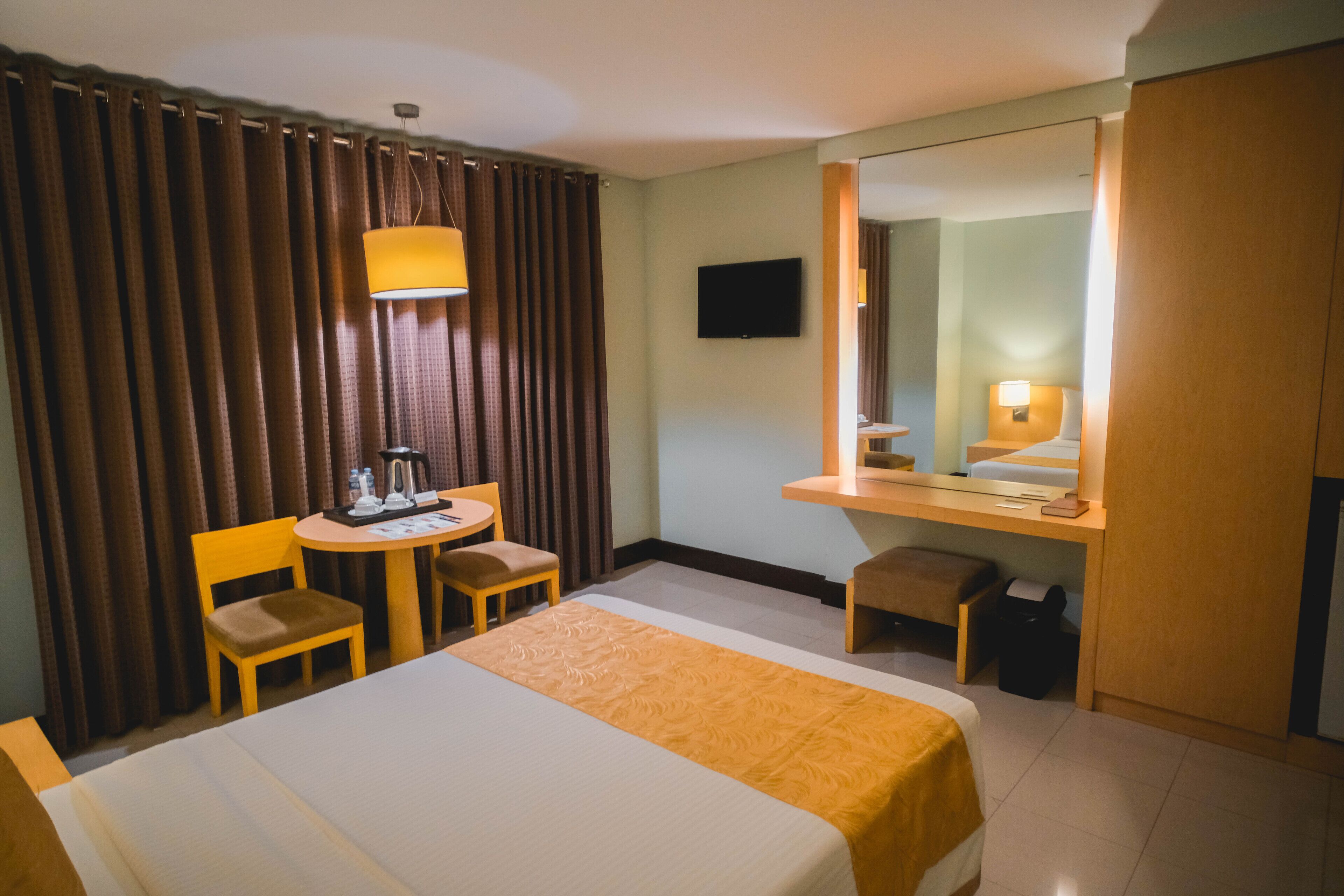 A hotel room with a bed, a desk with a chair, a small table, a mirror, and curtains.