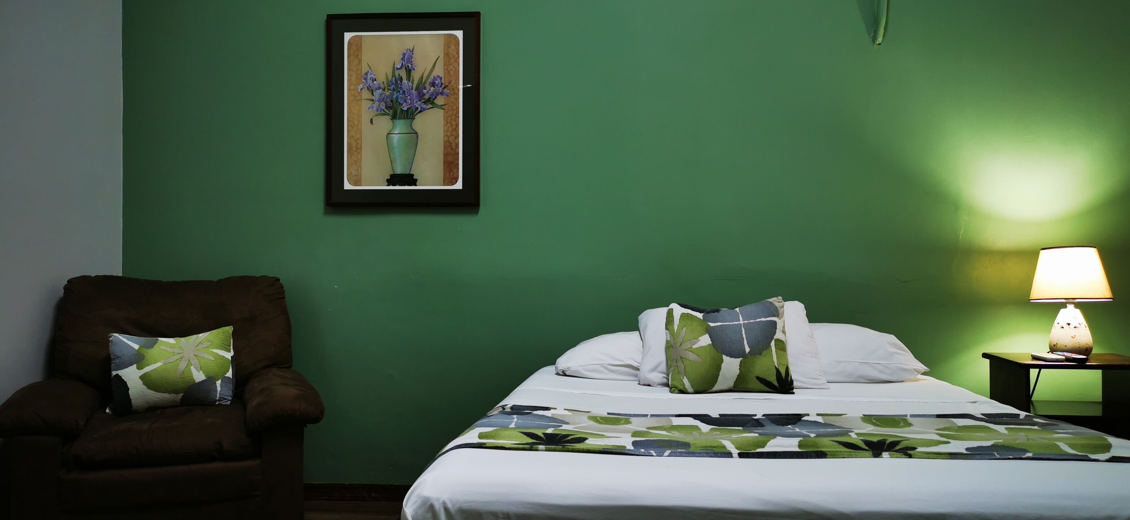 A room with a bed, a chair, a nightstand with a lamp, and a framed picture on the wall.
