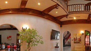 Un salón amplio con suelo de madera, un gran árbol en maceta, un tapete rojo, sofás de cuero y una escalera que sube a un nivel superior.