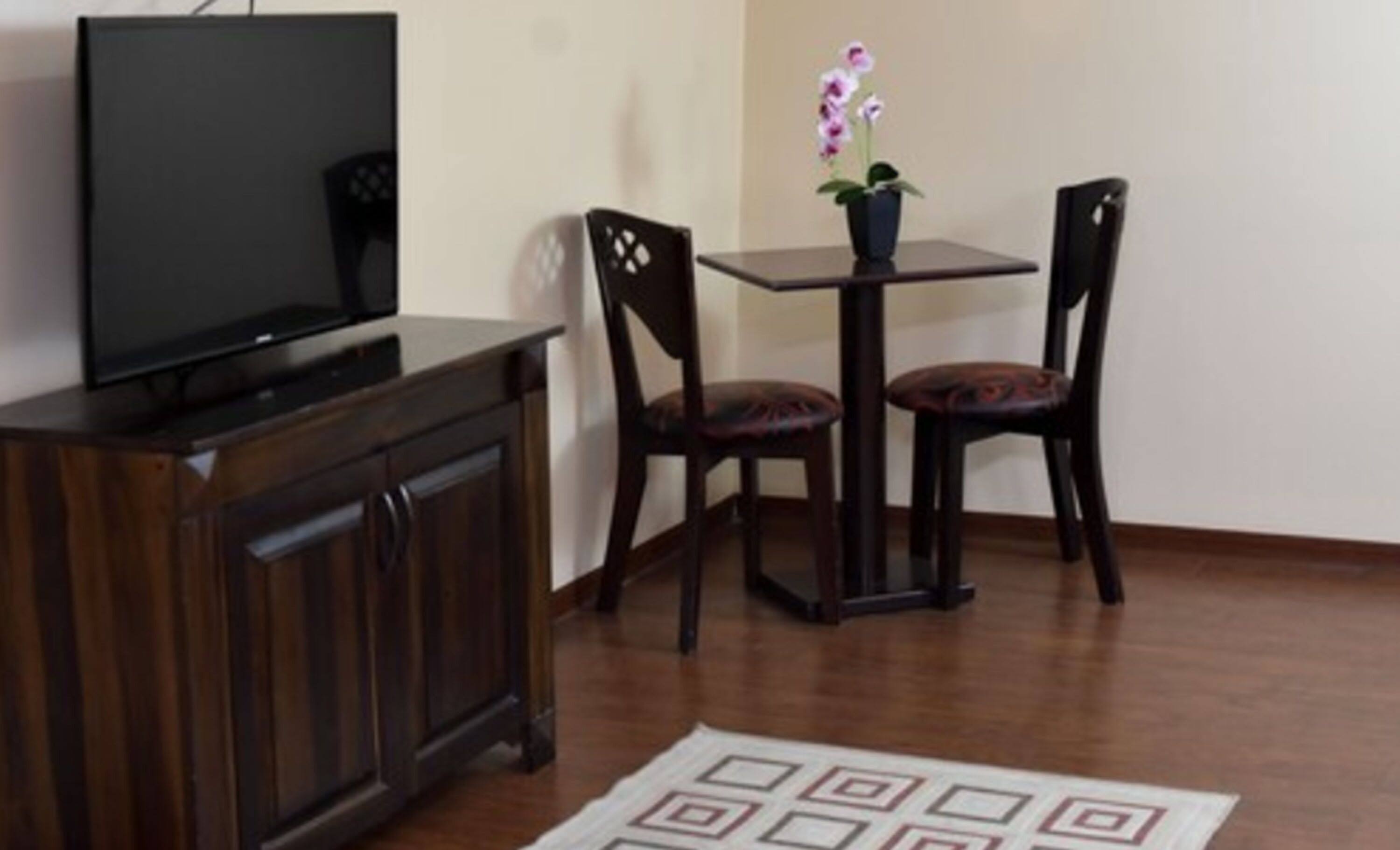 A room with a wooden cabinet, a flat-screen TV, two chairs, a small table, and a potted orchid.