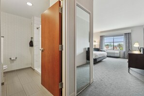 Habitación de hotel con puerta de madera, baño con inodoro y ducha, cama, escritorio y ventana con vistas a edificios.